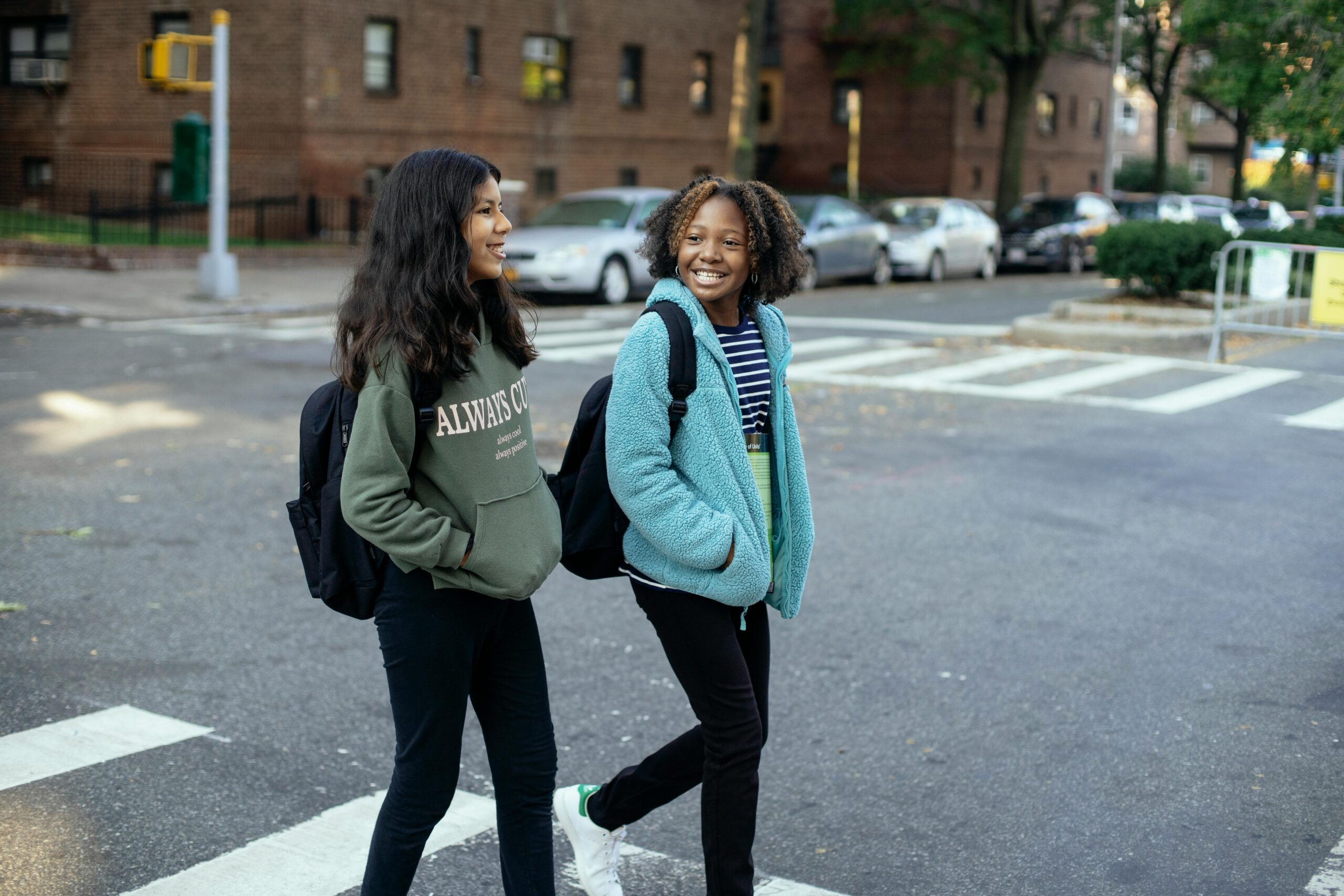 Two school-aged girls wearing back-packs smile as they walk across the crosswalk in a city.