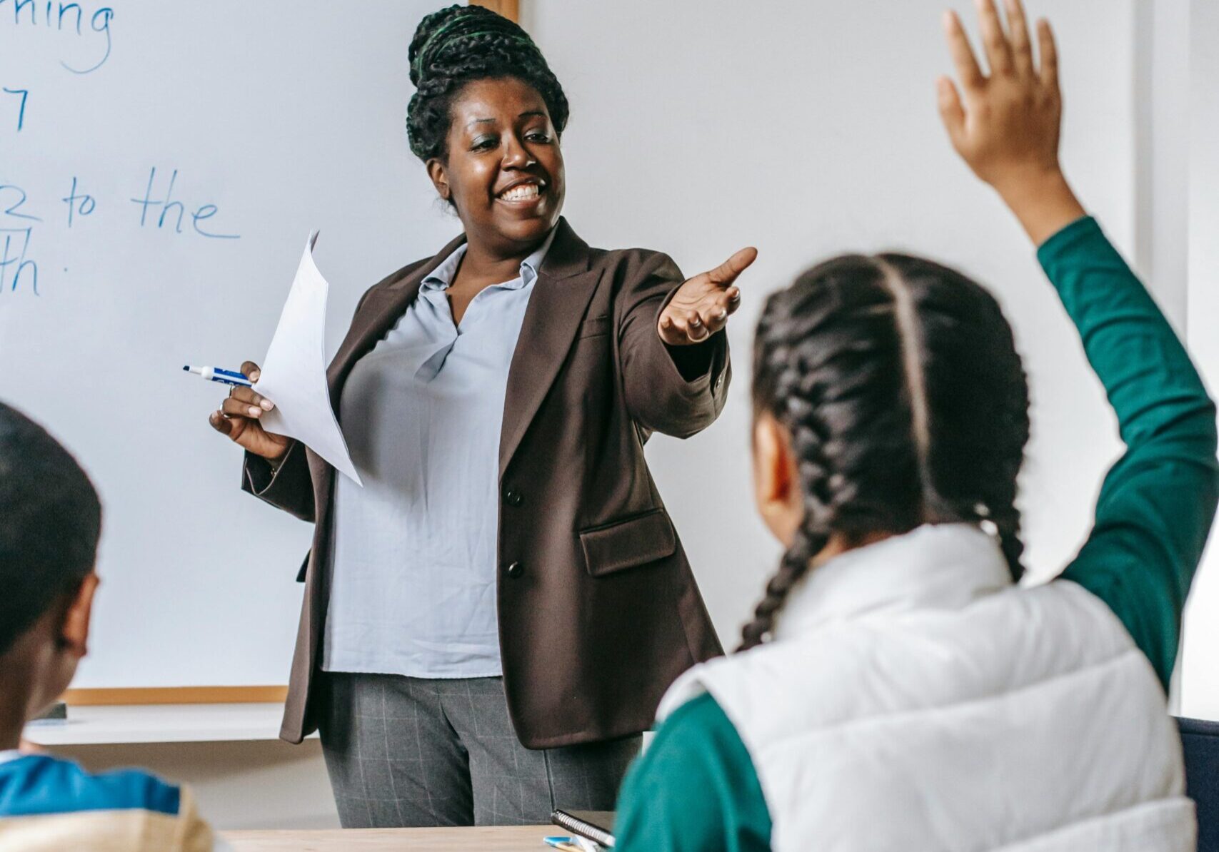 A smiling teacher calls on a student with a raised hand in classroom.